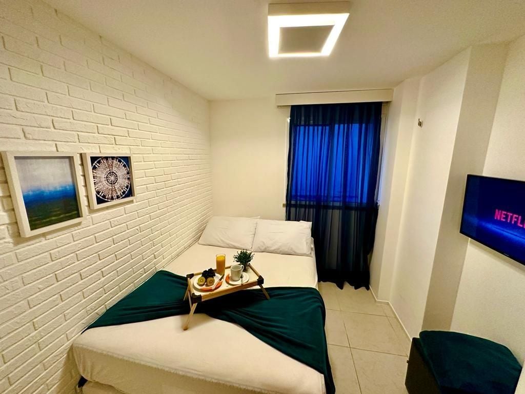 A hotel room with a white brick wall, a bed with a green bedspread, a tray with breakfast items, a TV displaying 'NETFLIX', and a window with blue curtains.