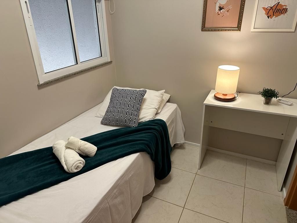 A small, neatly arranged bedroom with a bed, pillows, a bedside lamp, and a small desk with a lamp.