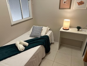 Quarto pequeno e bem arrumado, com cama, almofadas, abajur e uma escrivaninha com luminária.