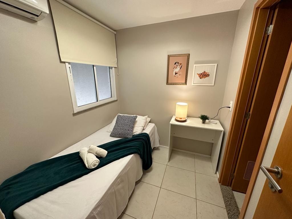 A small, neatly arranged bedroom with a bed, bedside table with a lamp, a window with blinds, and a door.
