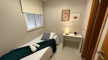 A small, neatly arranged bedroom with a bed, bedside table with a lamp, a window with blinds, and a door.