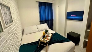 A hotel room with a bed, a TV mounted on the wall, a window with blue curtains, and a tray with breakfast items on the bed. - Porto Beach Conforto e Lazer a 200m do B Park (Aquiraz)