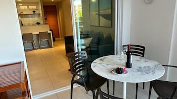 A dining area with a round table, black chairs, and a glass top. There is a plate with grapes and a bottle of wine. A kitchen is visible in the background.