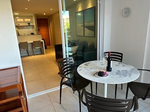 Uma área de refeições com uma mesa redonda, cadeiras pretas e tampo de vidro. Há um prato com uvas e uma garrafa de vinho. Uma cozinha é visível ao fundo.