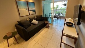 A living room with a sofa, a small table, a TV, and a balcony with chairs. - Porto Beach Conforto e Lazer a 200m do B Park (Aquiraz)