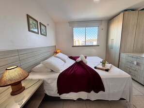 Una cama bien hecha con ropa de cama blanca, una manta roja y una pequeña planta en maceta sobre la mesita de noche.