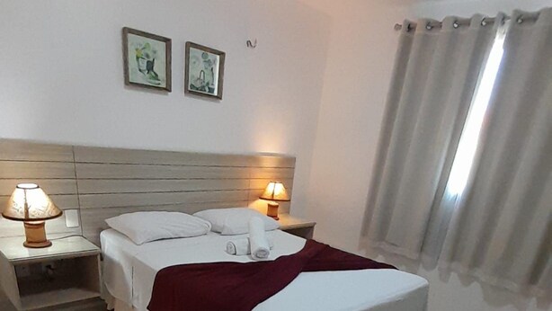 A neatly made bed with white linens and a red blanket, two bedside lamps, and two framed pictures on the wall. - Porto Beach Conforto e Lazer a 200m do B Park (Aquiraz)