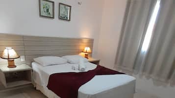 Una cama bien hecha con sábanas blancas y una colcha roja, dos lámparas de noche y dos cuadros en la pared.