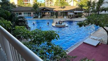 A swimming pool with a circular hot tub, surrounded by palm trees and a building with balconies.