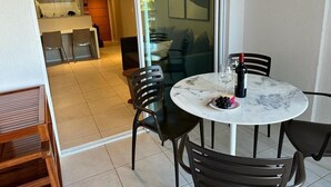 A dining area with a round table, black chairs, and a glass top. There is a plate with grapes and a bottle of wine. A kitchen is visible in the background. - Porto Beach Conforto e Lazer a 200m do B Park (Aquiraz)