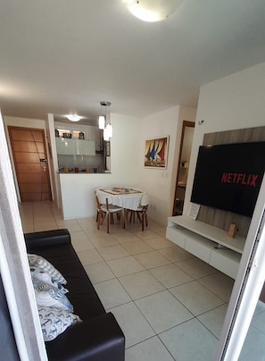 A living room with a sofa, a dining table with chairs, a kitchen area, and a TV on a stand. - Porto Beach Conforto e Lazer a 200m do B Park (Aquiraz)