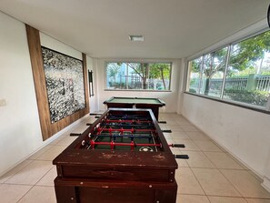 A room with a foosball table and a pool table, large windows with a view of greenery, and a wall-mounted artwork. - Porto Beach Conforto e Lazer a 200m do B Park (Aquiraz)