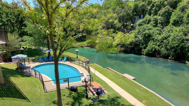 Ein Schwimmbecken, umgeben von einer Sonnenterrasse mit Liegen und Sonnenschirmen, ein kleiner Terrassenbereich und ein Fluss.