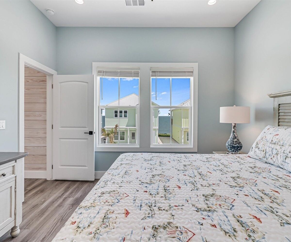 A bedroom with a large bed, a nightstand, a lamp, and a view of a green house through a window.