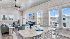 A spacious living room with a large wooden dining table, comfortable seating, and large windows offering views of the surrounding area. - Sunshine And Flip Flops Sp114 4 Bedroom Home by RedAwning (Fulton)