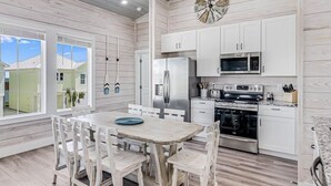 A modern kitchen with white cabinetry, stainless steel appliances, and a wooden dining table with chairs. - Sunshine And Flip Flops Sp114 4 Bedroom Home by RedAwning (Fulton)