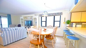A spacious living area with a striped sofa, a dining table with chairs, a TV, and a kitchen area with stools. - Amber Waves 300 3 Bedroom Condo by RedAwning (Ocean City)