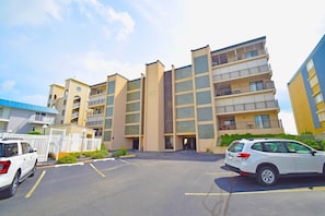 A multi-story residential building with a central entrance, parking lot, and a gated driveway.