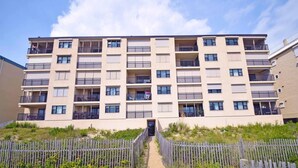A multi-story residential building with balconies, a gated entrance, and a sandy pathway leading to the entrance. - Amber Waves 300 3 Bedroom Condo by RedAwning (Ocean City)