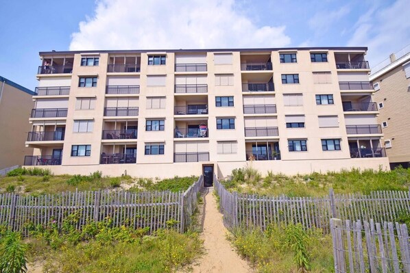 A multi-story residential building with balconies, a gated entrance, and a sandy pathway leading to the entrance.