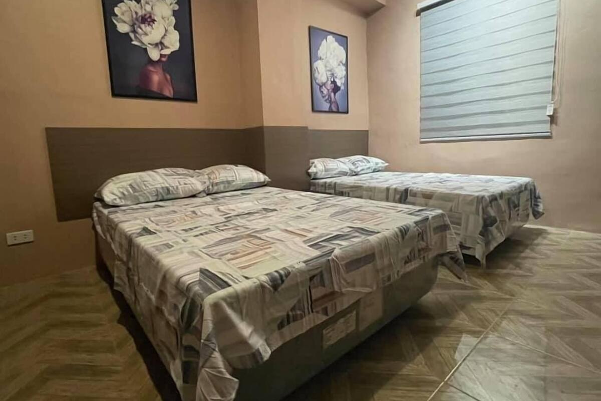A bedroom with two beds, a large window with blinds, and floral artwork on the walls.