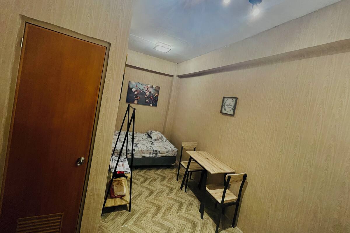 A bedroom with a bed, a desk, and a chair. There is a door, a painting on the wall, and a ceiling light fixture.