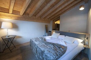 Quarto com teto de madeira, cama com roupa de cama branca, mesa de cabeceira com abajur e pequena mesa redonda também com abajur.