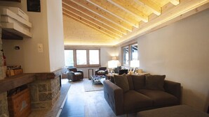 A spacious living room with a stone fireplace, wooden ceiling beams, and a large window providing natural light.