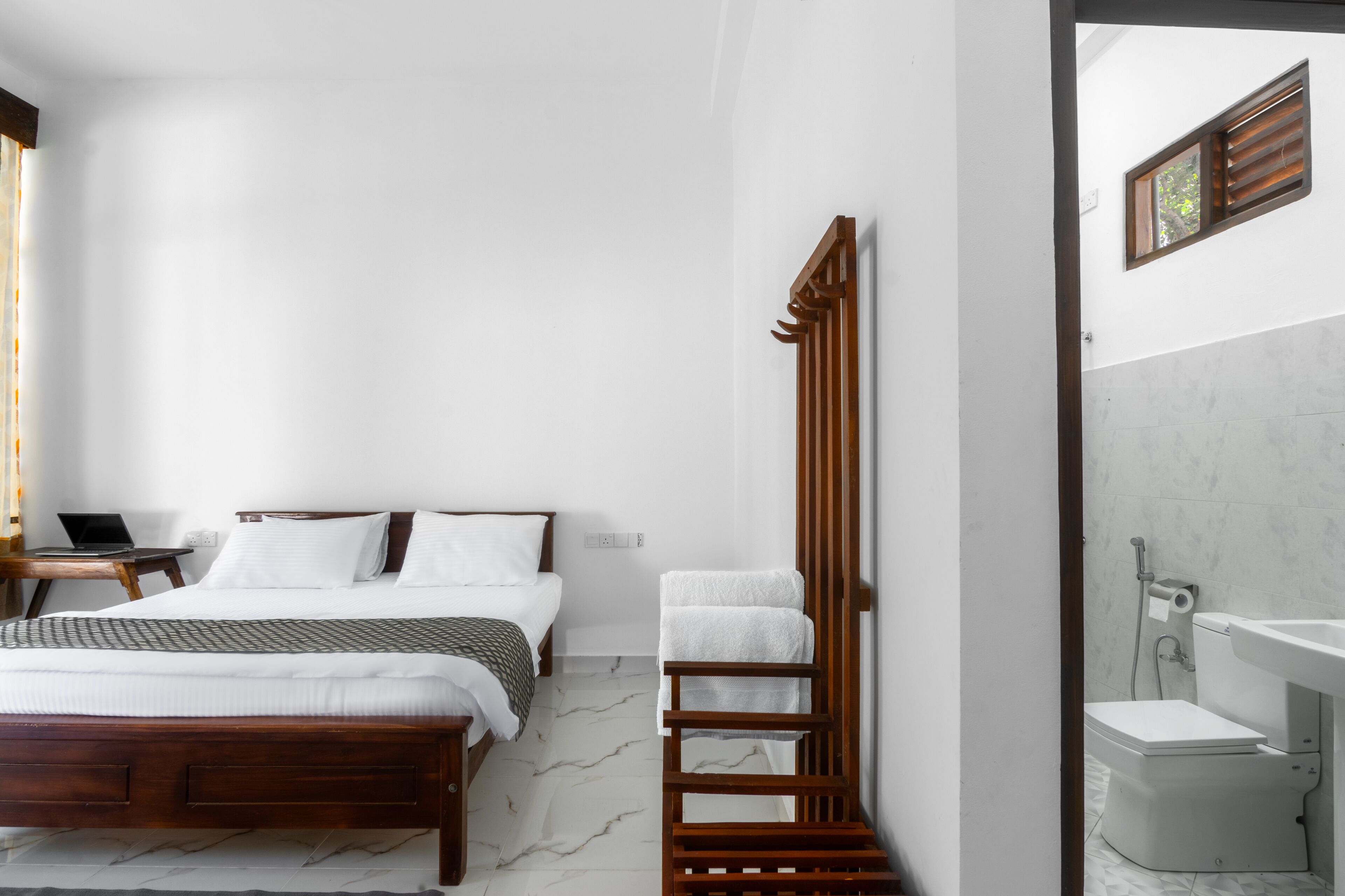Un dormitorio con una cama de madera, una mesita de noche con una computadora portátil y un baño con inodoro y lavamanos.