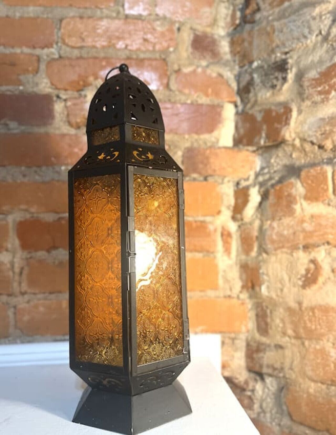Un farol con diseños elaborados y una base negra, colocado frente a un muro de ladrillo.