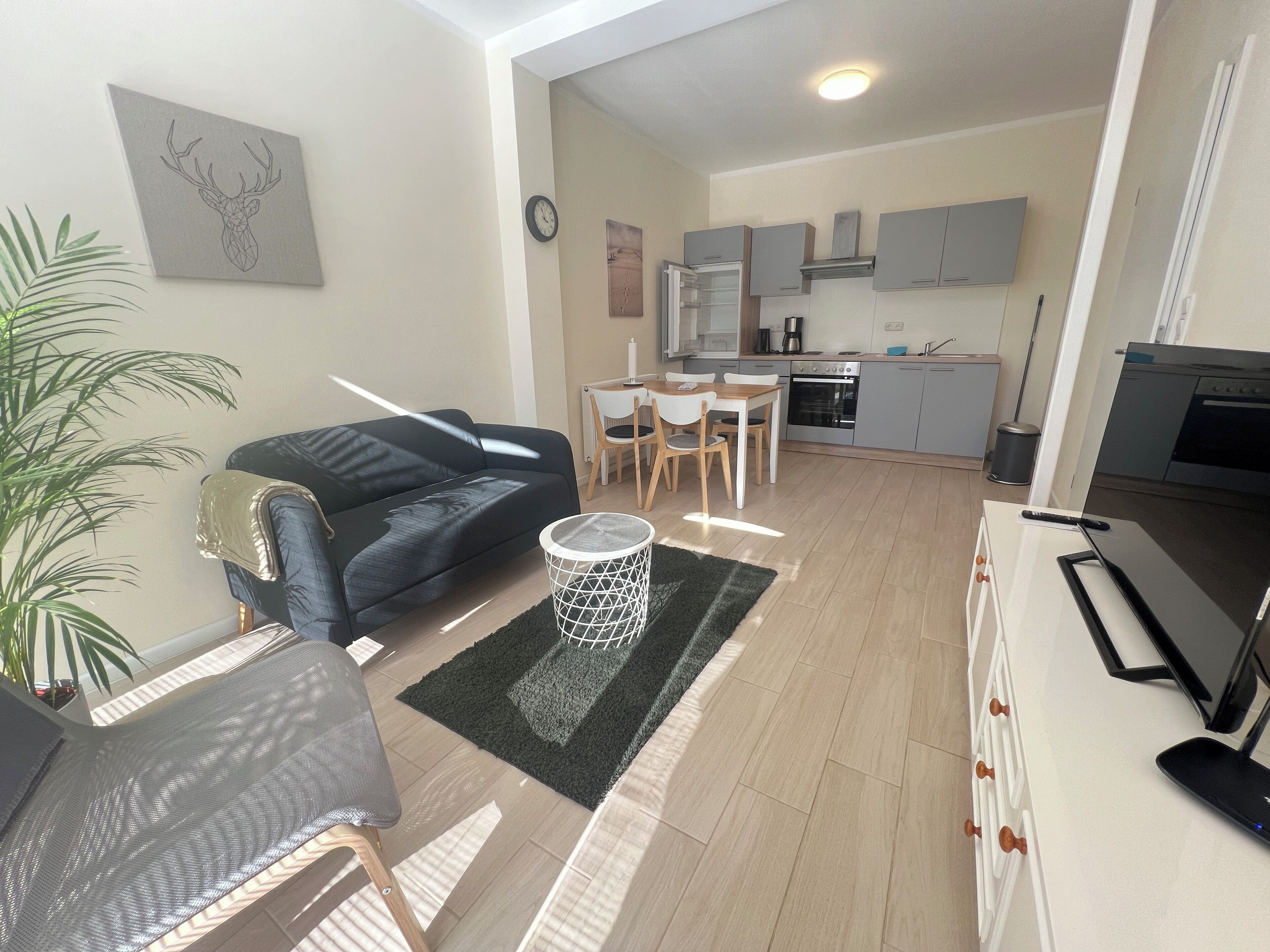 Ein modernes Wohnzimmer mit einer schwarzen Couch, einem Esstisch mit Stühlen, einer Küchenzeile mit Schränken und Geräten sowie einem Fernseher auf einem Schrank.