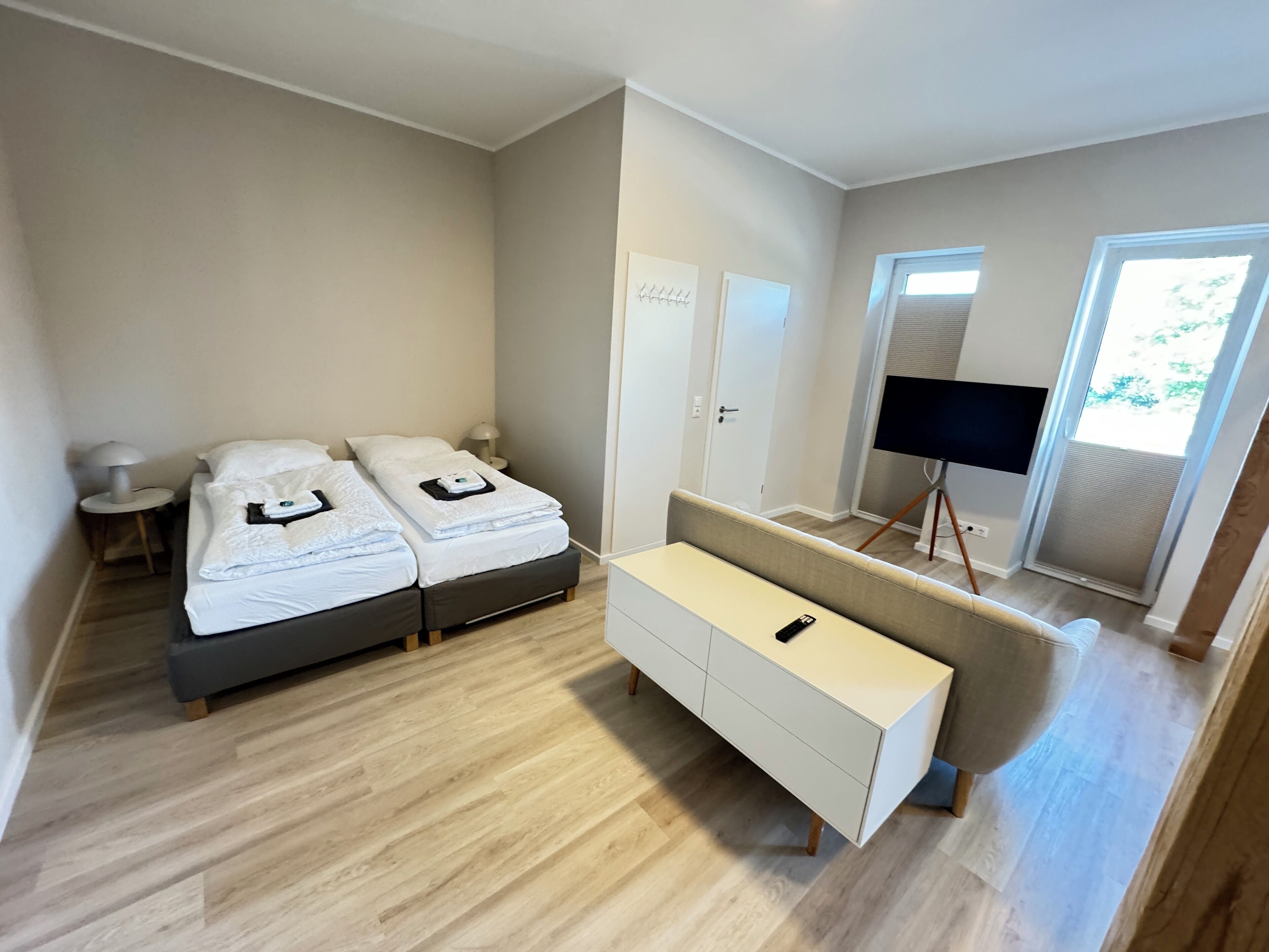 A modern hotel room with two beds, a sofa, a TV on a stand, and a door leading to a balcony.