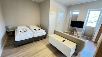 A modern hotel room with two beds, a sofa, a TV on a stand, and a door leading to a balcony.