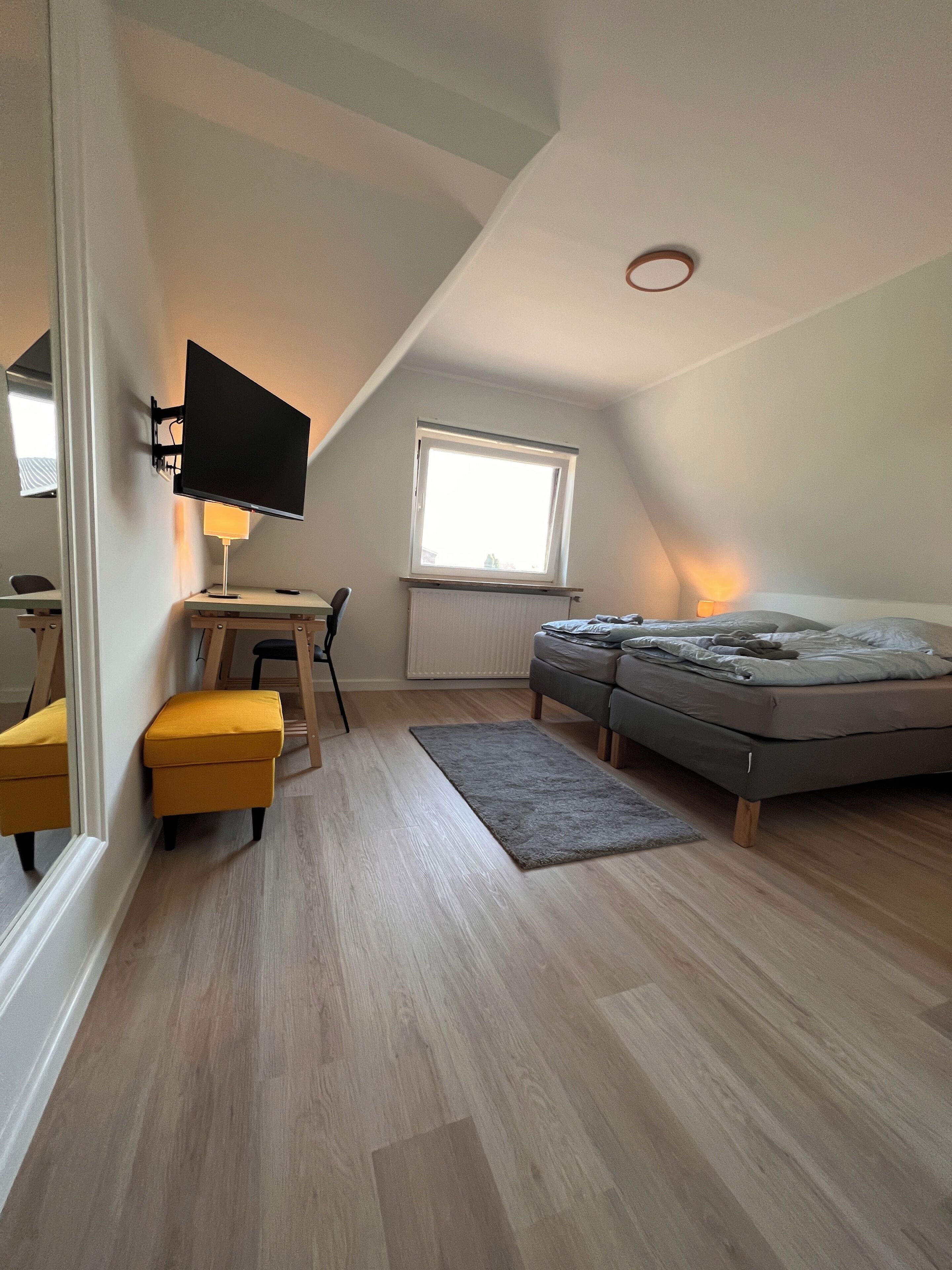 Een slaapkamer met een bed, een bureau met stoel, een televisie, een raam en een radiator.