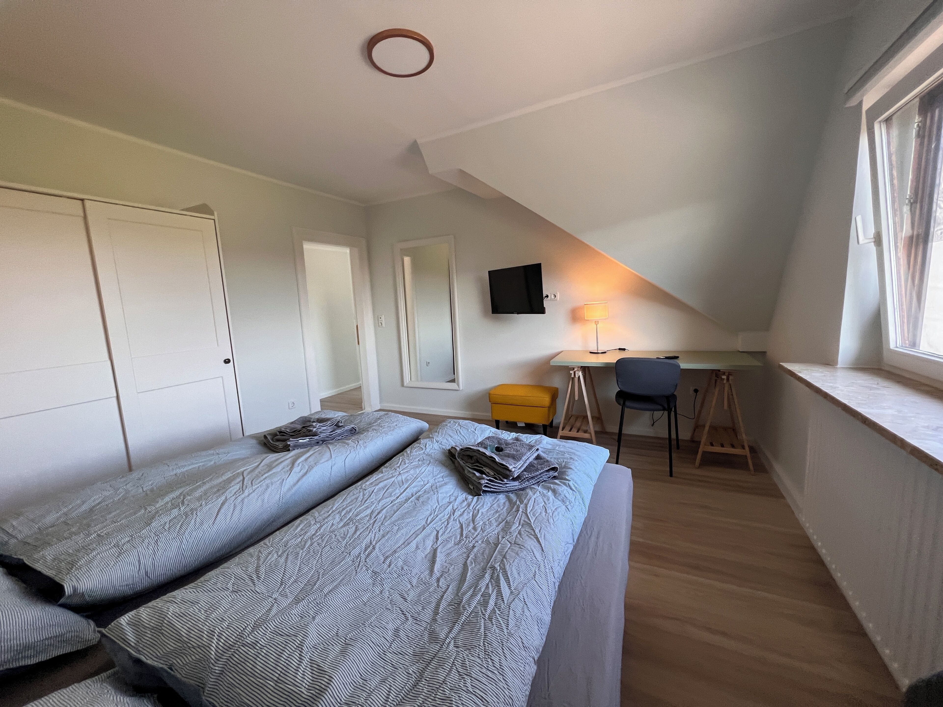 A dormitory room with two beds, a desk, a chair, a TV, and a window.