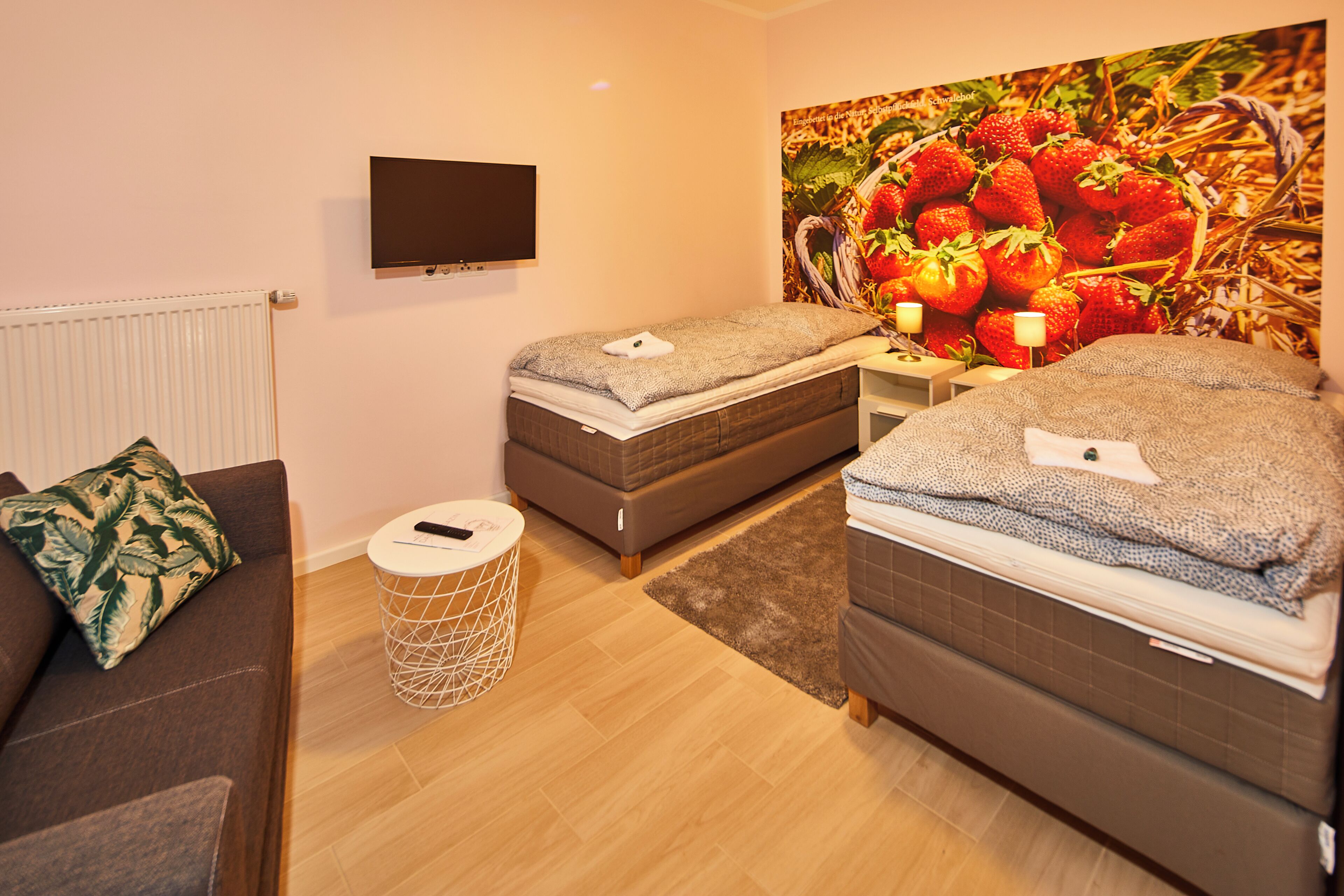 A hotel room with two beds, a TV, a large fruit-themed wall, a small round table, and a sofa.