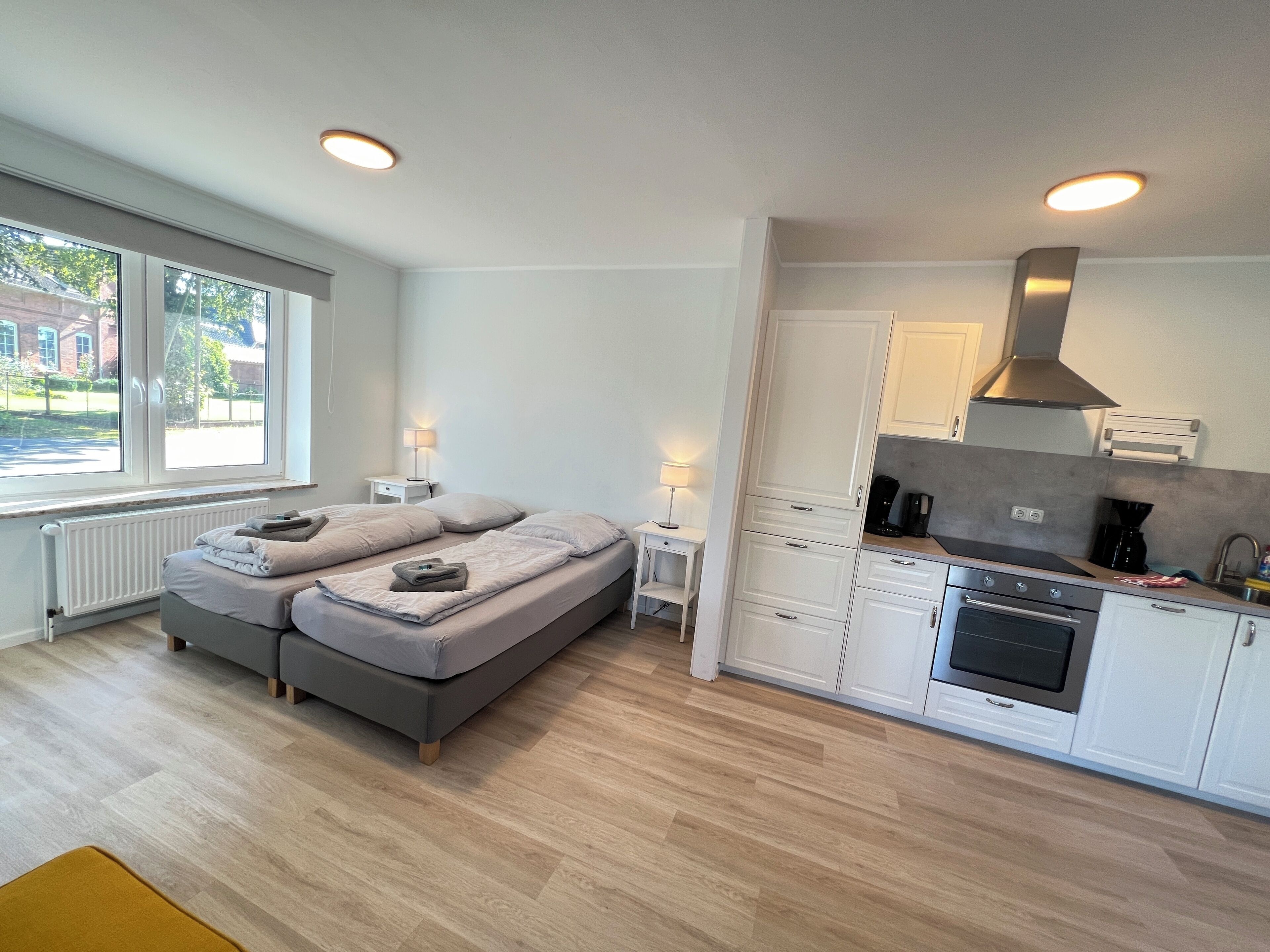 A modern room with a bed, bedside tables, a lamp, a kitchen with a stove, and a window with a view of the outside.