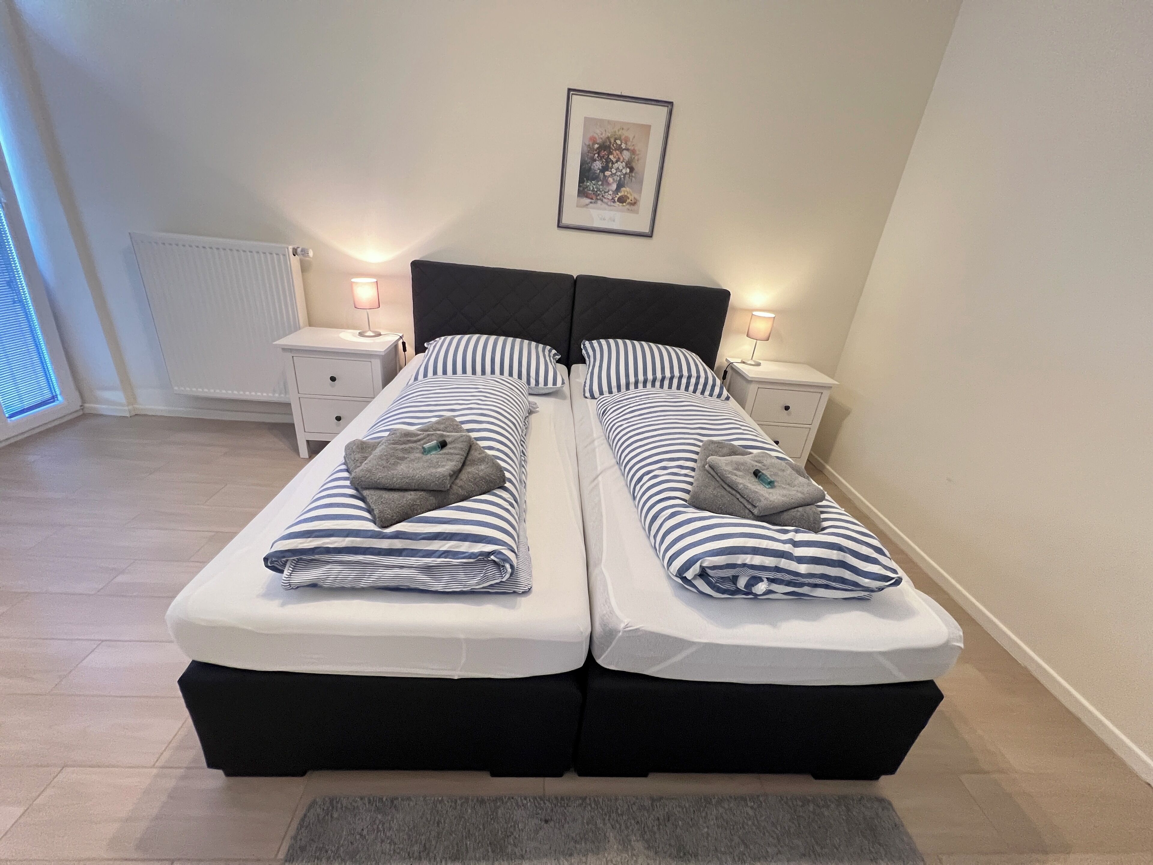 A double bed with striped bedding, two bedside tables with lamps, a framed picture on the wall, and a window with blinds.