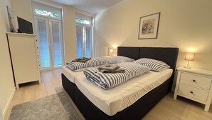 Une chambre à coucher avec un grand lit, une table de chevet, une lampe, un tableau encadré au mur et une fenêtre avec des stores.