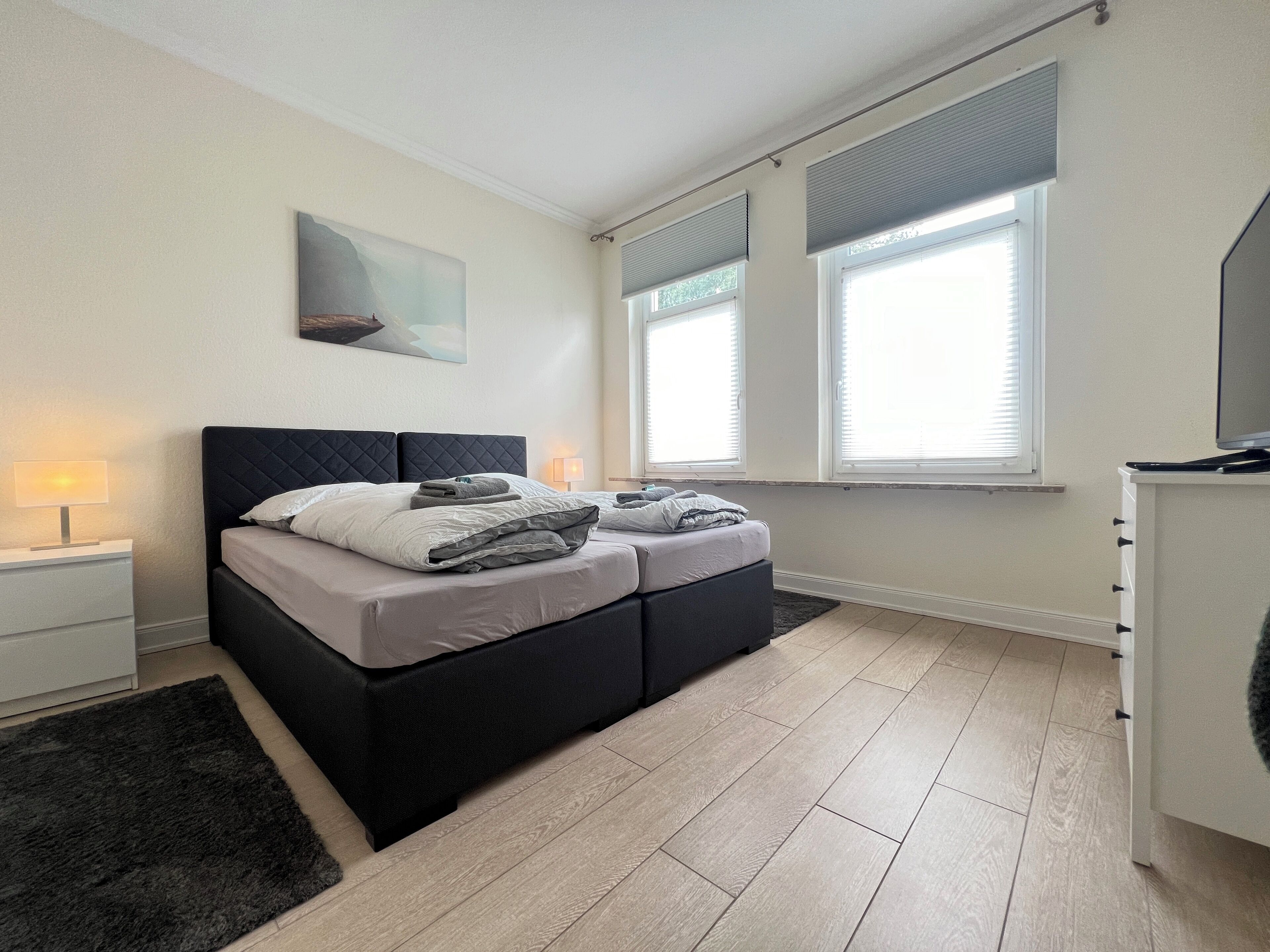 Een moderne slaapkamer met een bed, nachtlampjes, een klein vloerkleed en een raam met luxaflex.