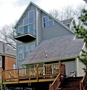 Une maison à deux étages avec un balcon à l’étage supérieur et une terrasse en bois avec un escalier menant à l’entrée.