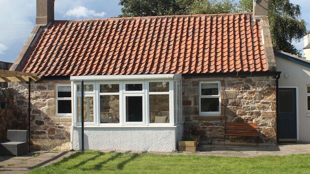 Une maison en pierre avec un toit en tuiles, un porche couvert avec des fenêtres et un banc à l’extérieur.