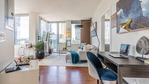 A modern living room with a sofa, a desk with a laptop, a coffee table, and a large window offering a city view. - 2BR | Gym, Pool | Bellevue | Sophari Towers M (Bellevue)