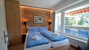 Un lit double avec une couette à carreaux bleus, une tête de lit en bois, un tableau encadré au-dessus du lit et une vue sur un espace extérieur aménagé, visible à travers une grande fenêtre.