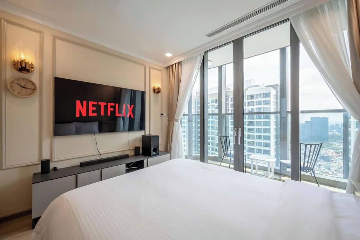 Ein Hotelzimmer mit einem großen Bett, einem an der Wand befestigten Fernseher, der das Netflix-Logo zeigt, einem Schreibtisch mit einem Computer und Blick auf die Stadt durch große Fenster.