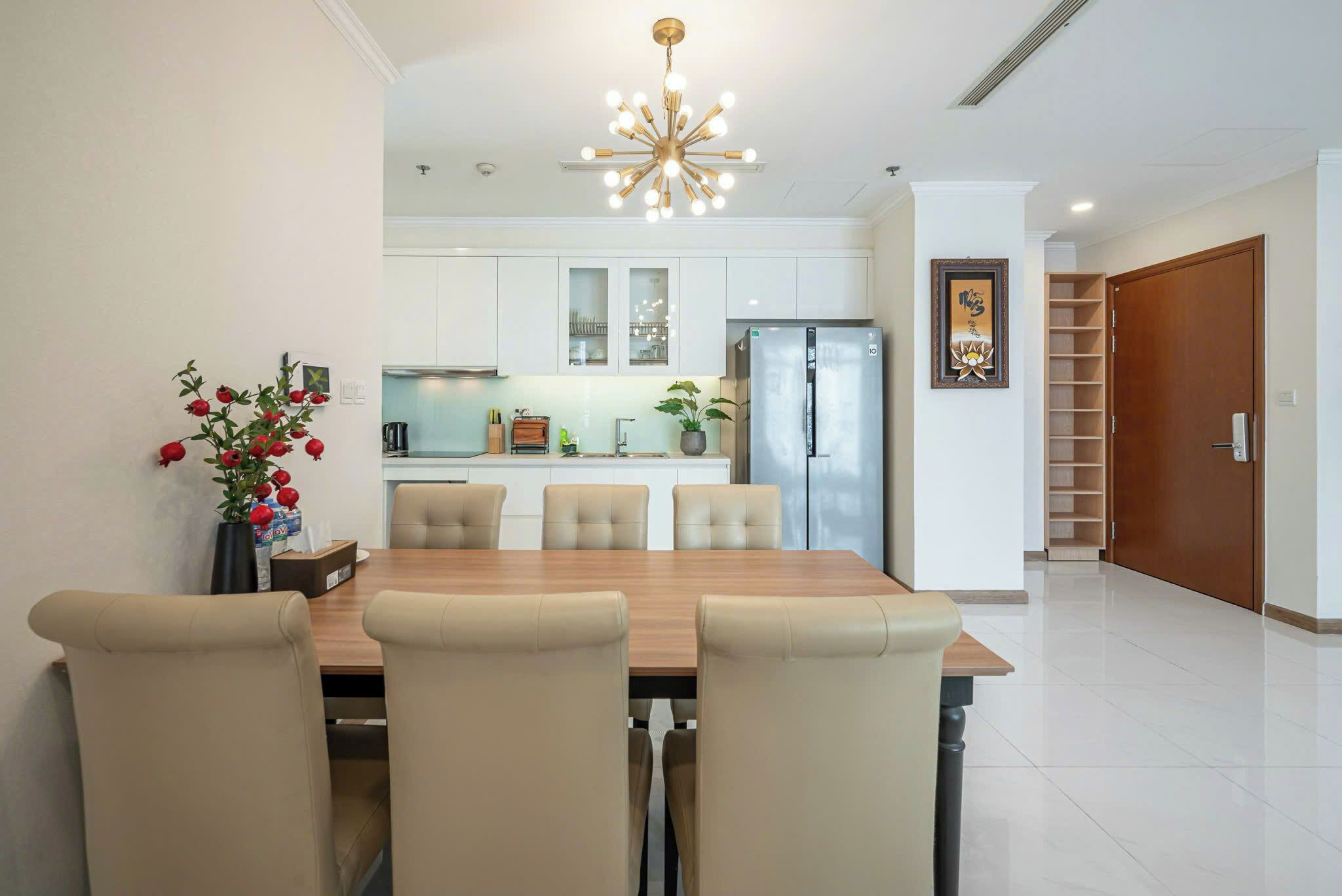 A modern kitchen with white cabinets, a stainless steel refrigerator, and a dining area with a wooden table and beige chairs.