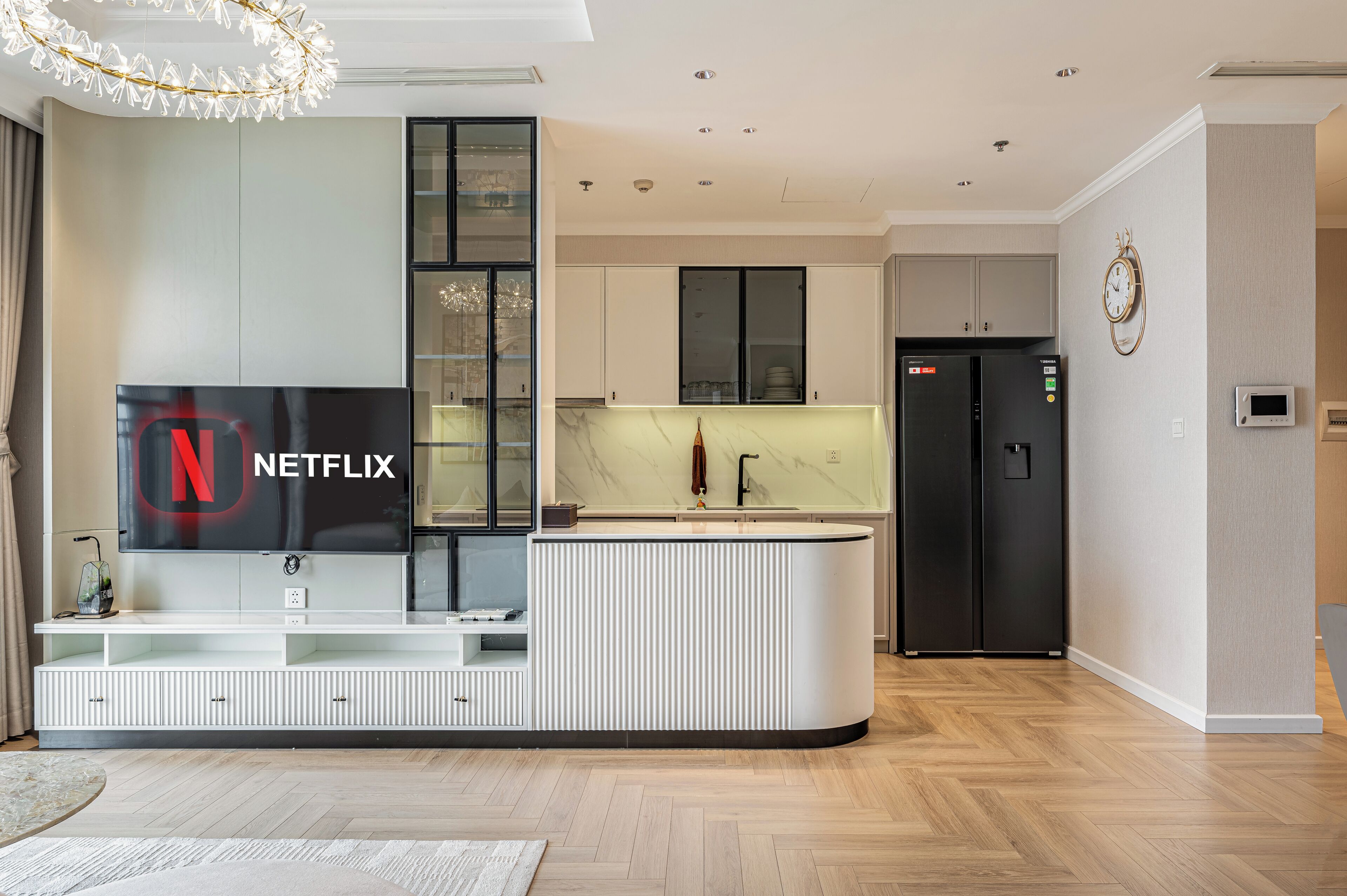 Ein modernes Wohnzimmer mit integriertem Fernseher, einer Küchenzeile mit Kühlschrank und einem Essbereich mit Tisch und Stühlen.