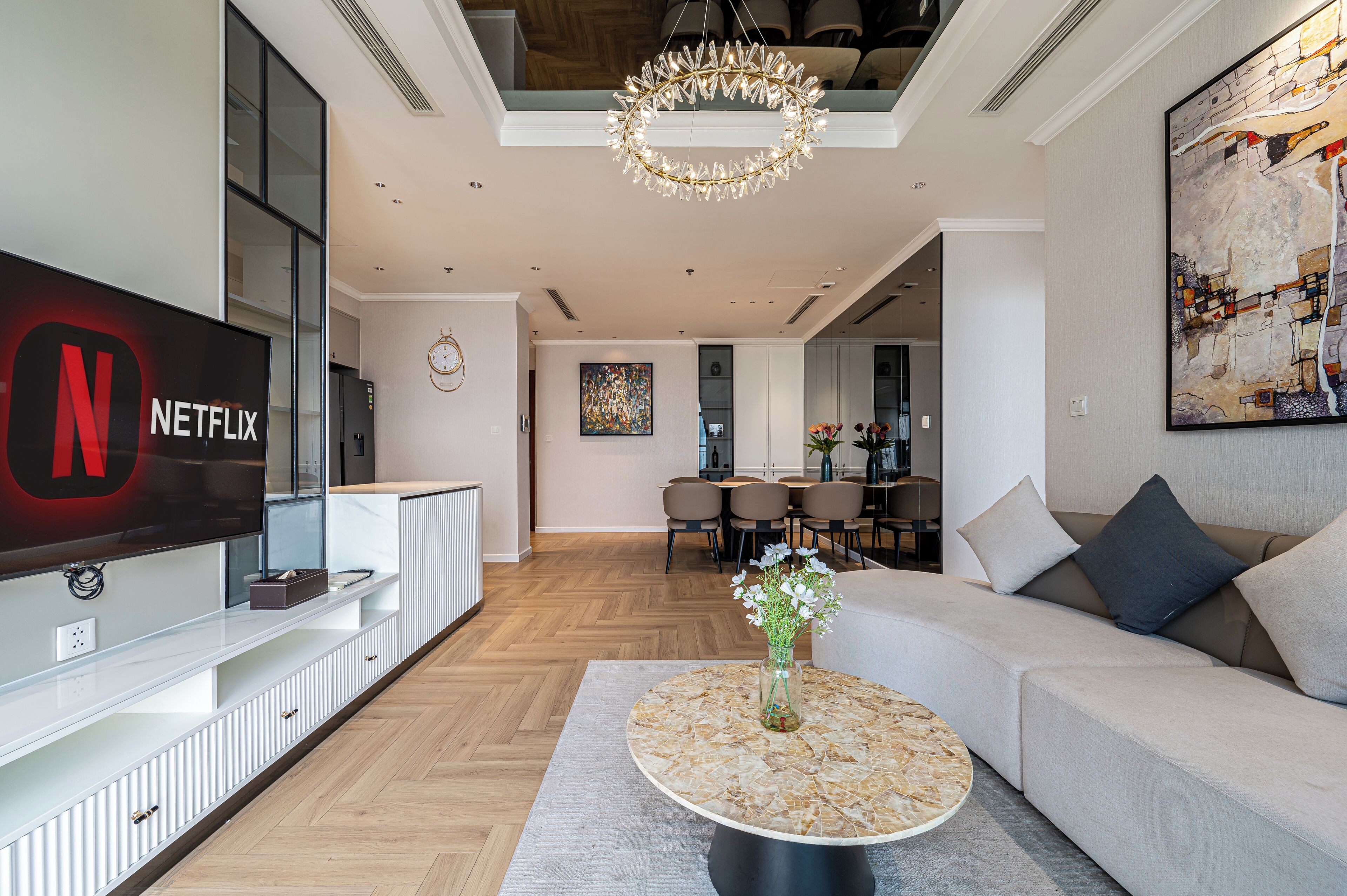 A modern living room with a flat-screen TV, a round coffee table with a vase of flowers, a sofa, and a dining area in the background.