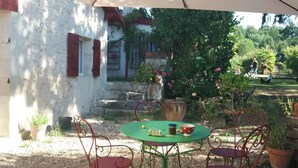 An outdoor patio with a table and chairs, surrounded by potted plants and a white building with red shutters.