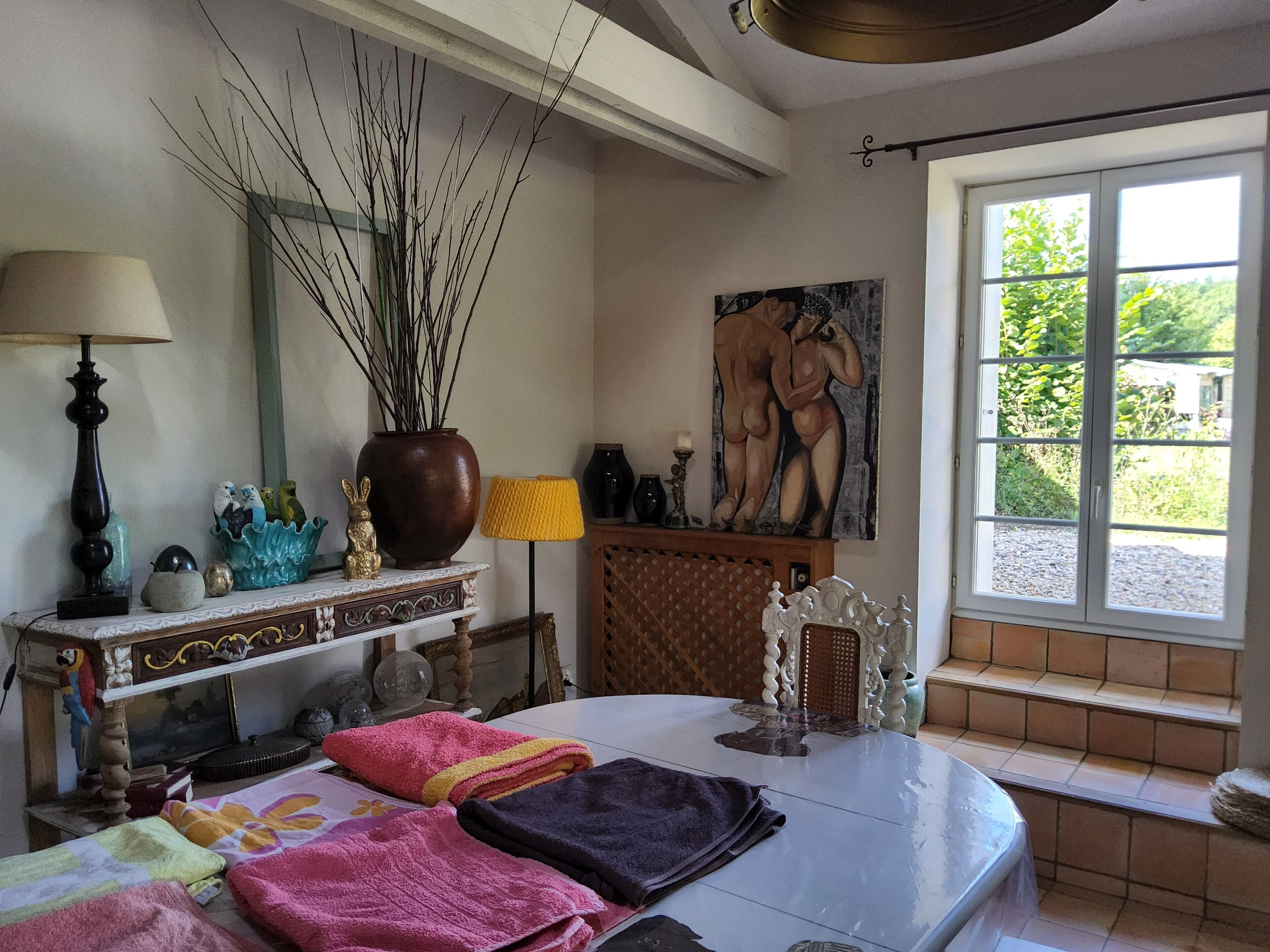 Una habitación luminosa con una cama, una mesita de noche con una lámpara, un jarrón con ramas, un cuadro y una ventana con vistas a zonas verdes.